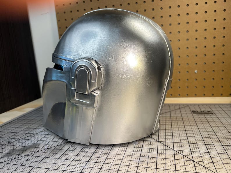 May include: A metallic silver helmet featuring a smooth, rounded dome and a distinct T-shaped visor opening. The side of the helmet displays a raised, rectangular ear piece detail with a vertical slot, finished in a uniform, reflective silver paint.