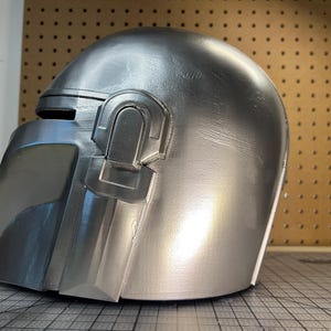 May include: A metallic silver helmet featuring a smooth, rounded dome, a T-shaped visor opening, and a distinct raised rectangular earpiece detail on the side. The surface has a reflective finish with subtle texture visible across the curved exterior.