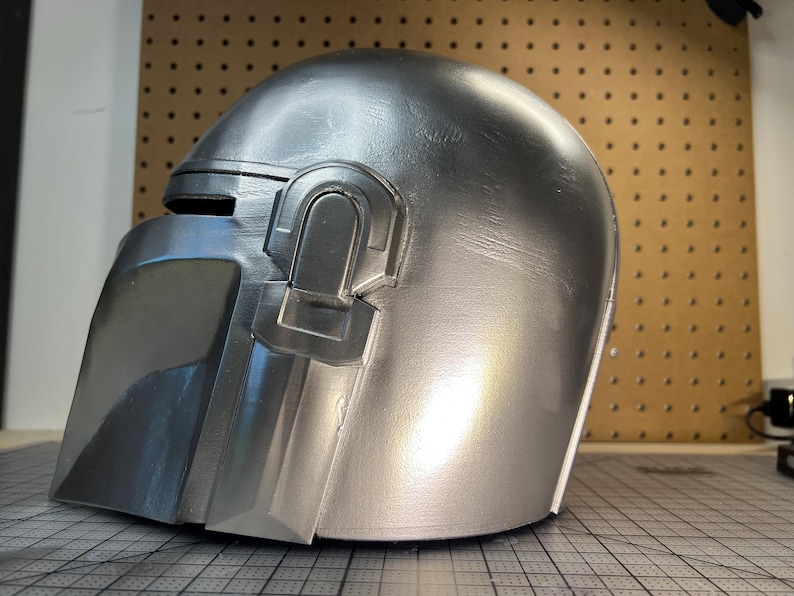 May include: A metallic silver helmet featuring a smooth, rounded dome, a T-shaped visor opening, and a distinct raised rectangular earpiece detail on the side. The surface has a reflective finish with subtle texture visible across the curved exterior.