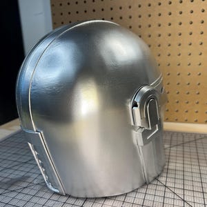 May include: A smooth, metallic silver helmet featuring a rounded dome shape, a distinct T-shaped visor opening, and raised rectangular detailing on the side. The surface has a reflective finish with visible seams along the construction of the piece.