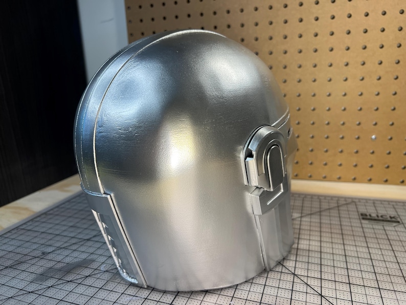 May include: A smooth, metallic silver helmet featuring a rounded dome shape, a distinct T-shaped visor opening, and raised rectangular detailing on the side. The surface has a reflective finish with visible seams along the construction of the piece.