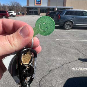 Puede incluir: Un llavero verde con una cara sonriente se sostiene frente a un estacionamiento y una tienda Aldi. El llavero está sujeto a un mosquetón negro con llaves y un llavero de coche.