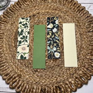 Puede incluir: Cuatro marcadores rectangulares sobre una estera tejida. Un marcador presenta un fondo crema con hojas verdes y una flor rosa. Otro es verde liso, y un tercero tiene un fondo negro con patrones florales blancos y azules. El último es de color crema liso.
