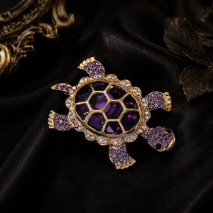 Könnte beinhalten: Goldfarbene Schildkrötenbrosche mit einem lila Edelsteinpanzer und lila und klaren Kristallakzenten. Die Brosche ist vor einem schwarzen Hintergrund platziert, der ihr aufwendiges Design und ihre funkelnden Details hervorhebt. Ein dekoratives Accessoire.