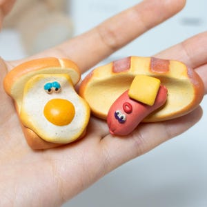 Könnte beinhalten: Zwei Miniatur-Lebensmittelskulpturen: eine Toastscheibe mit Spiegelei und ein Hotdog im Brötchen. Das Ei hat ein gelbes Eigelb und blaue Augen. Der Hotdog hat ein gelbes Butterquadrat und blaue Augen. Die Artikel sind in einer Hand.