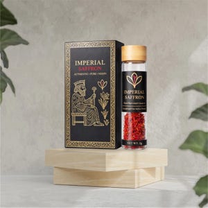 May include: A black box and a glass jar of Imperial Saffron sit on a wooden display. The box features gold text and an illustration of a king. The jar has a wooden lid and contains red saffron threads. The text on the jar reads "NET WT. 2g".