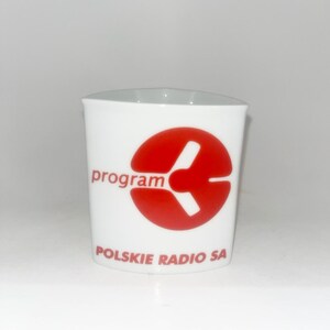 May include: A white ceramic mug with a slightly triangular rim. The front features a red circular logo with a stylized white graphic inside, the word "program" in red text, and "POLSKIE RADIO SA" printed in red at the bottom.