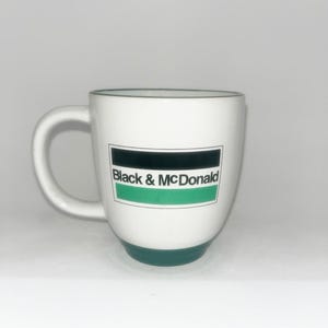 May include: A white ceramic mug featuring a handle on the left side and a rectangular logo on the front. The logo contains a black horizontal bar above the text Black & McDonald, which sits above a green horizontal bar. The base is dark green.