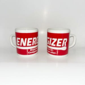 May include: Two white ceramic mugs with red bands featuring the Energizer logo in white text. The left mug displays the word ENERGIZER and the text Depend on it! Compter sur elle!. The right mug displays the word ENERGIZER and the text Depend on it!