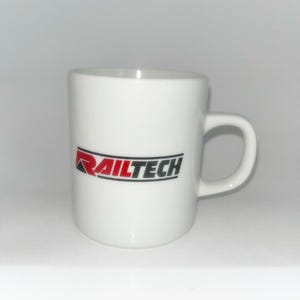 May include: A white ceramic mug featuring the text RAILTECH printed in a bold, stylised font. The word RAIL is in red, while the word TECH is in black, with a black horizontal line running through the centre of the text on the front side.