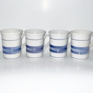 May include: A set of four white ceramic mugs, each featuring a series of horizontal blue stripes in varying shades of light and dark blue. The mugs have a cylindrical body, a flared rim, and a curved handle on the side for holding the vessel.