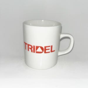 May include: A white ceramic mug featuring the word TRIDEL printed in bold red capital letters on the front. The letter D in the word contains a small white triangle cutout. The mug has a smooth finish, a cylindrical body, and a curved handle.
