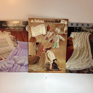 May include: Three vintage craft instruction booklets featuring One Stitch At A Time with Ellen McCarn, a Patons baby knitting collection, and a Bernat pattern book. The covers display various knitted garments, baby clothes, and home decor items.