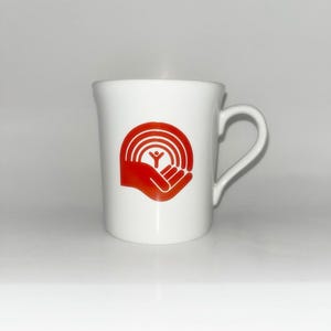 May include: A white ceramic mug featuring a red graphic logo on the front. The logo depicts a stylised hand cradling a figure beneath a series of concentric semicircles. The mug has a smooth finish, a cylindrical body, and a curved handle on the right side.