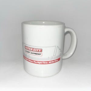 May include: A white ceramic mug featuring the Inter-City Fleet Express logo in red and black text. Below the logo, a red horizontal stripe contains the white text: The Right Place. The Right Time. All the Time. The mug has a curved handle on the side.