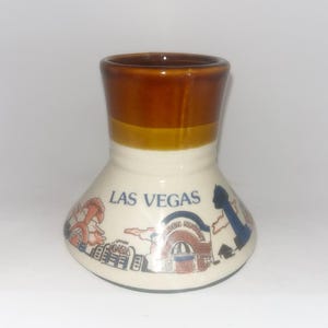 May include: A ceramic souvenir shot glass with a flared base and a brown glazed rim. The white body features a printed graphic of Las Vegas landmarks, including the Golden Nugget and Union Plaza, with the text LAS VEGAS printed in blue.