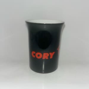 Peut inclure: Un mug en céramique noire avec un intérieur blanc, portant le nom CORY imprimé en lettres capitales rouges sur le devant. Le mug présente un bord légèrement évasé et une finition lisse et brillante, offrant une vue claire du texte et du contraste.