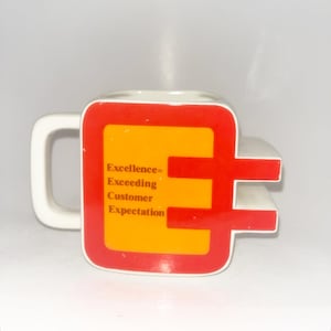 May include: A white ceramic mug with a square-shaped body featuring a red border and a yellow center panel. The text "Excellence= Exceeding Customer Expectation" is printed in brown on the yellow panel. The mug has a handle and a protruding side.
