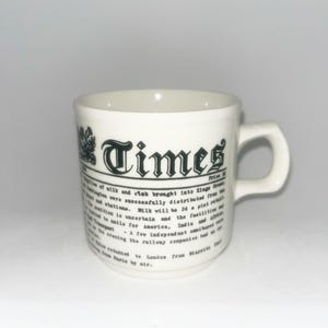 May include: A white ceramic mug featuring a black printed design that mimics a vintage newspaper front page. The text includes the word Times in a bold gothic font, followed by several columns of small, detailed news article text.