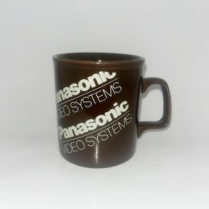 May include: A dark brown ceramic mug featuring the white text Panasonic VIDEO SYSTEMS printed repeatedly in a diagonal pattern across the front. The mug has a smooth finish, a cylindrical body, and a single curved handle on the right side.