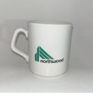 May include: A white ceramic mug featuring a green geometric logo and the text "northwood" in black lowercase letters. The mug has a smooth finish, a cylindrical body, and a curved handle on the left side, providing a simple and functional design.