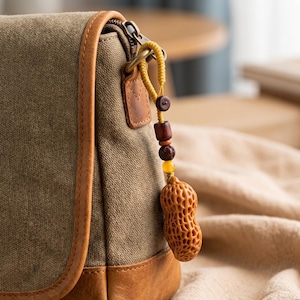 Könnte beinhalten: Nahaufnahme einer Canvas-Tasche mit braunem Lederbesatz und einem erdnussförmigen Anhänger. Der Anhänger ist aus geschnitztem Holz gefertigt und hängt an einer gelben Kordel mit Holz- und bernsteinfarbenen Perlen. Die Tasche ist olivgrün.