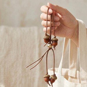 May include: A brown acorn pendant with four acorn charms. The acorn caps are a light brown color, and the acorn bodies are a darker brown. A gold-colored bead is at the center of the pendant.