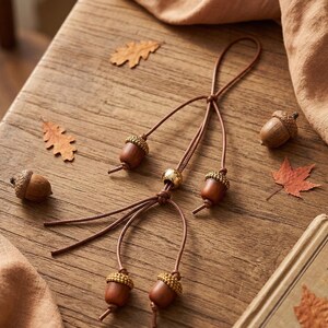 May include: A close-up of a wooden table with autumn-themed decorations. A brown leather cord with acorn pendants and a gold bead is the focal point. Scattered around are real acorns and dried leaves in shades of brown and orange.