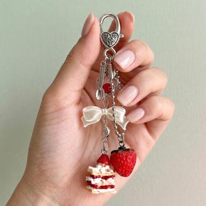 May include: A silver-coloured keyring with a heart-shaped clasp and hanging charms. Charms include a miniature spoon, a red heart, a white bow, a strawberry, and a layered cake with red filling. The keyring is held in a hand.