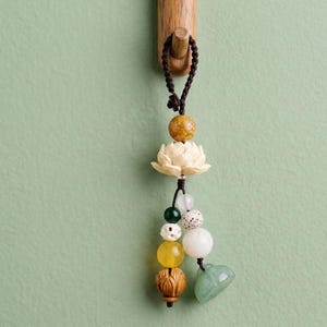 May include: A decorative beaded charm featuring a carved ivory lotus flower, various colorful beads, and a jade heart-shaped pendant. The charm is suspended from a dark brown cord and hangs from a wooden hook against a light green background.