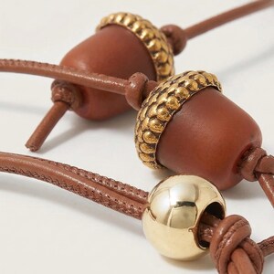 May include: Close-up of a brown leather necklace featuring acorn-shaped pendants with gold-colored caps. A gold sphere is also visible. The necklace has a knotted design.