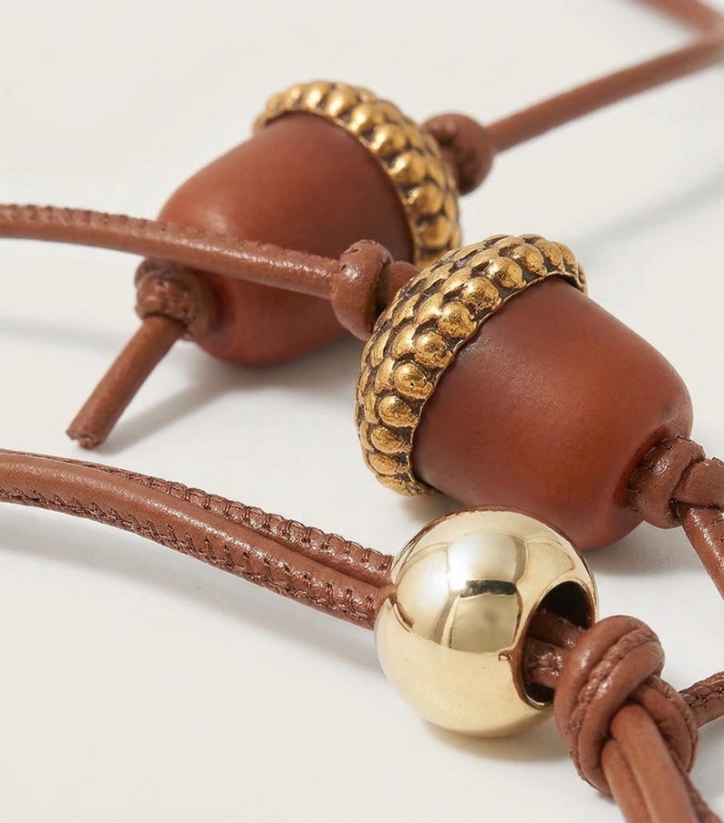 May include: Close-up of a brown leather necklace featuring acorn-shaped pendants with gold-colored caps. A gold sphere is also visible. The necklace has a knotted design.