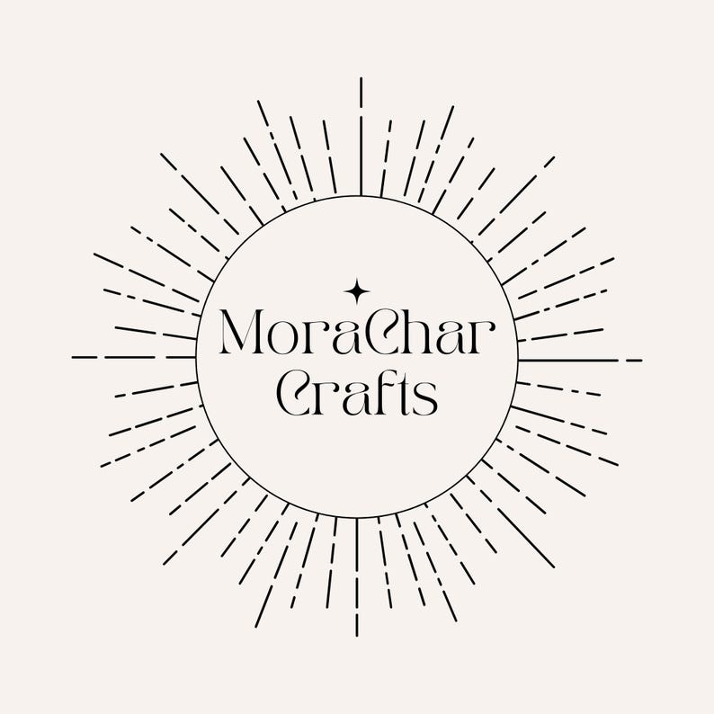 May include: A circular logo with the text "MoraChar Crafts" in a stylized font, surrounded by radiating lines. A small star is above the "a" in "Char". The design is in black on a cream-colored background.