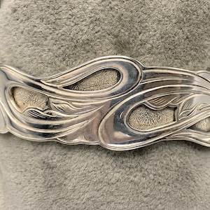 May include: A silver cuff bracelet with an Art Nouveau design. The bracelet features flowing, organic lines and textured, oval-shaped details. The metal has a polished finish, and the design is symmetrical.