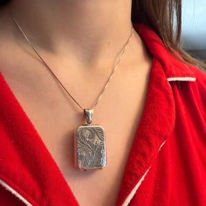 May include: A silver rectangular pendant necklace with an ornate floral design. The pendant hangs from a delicate silver chain. The person is wearing a red top.