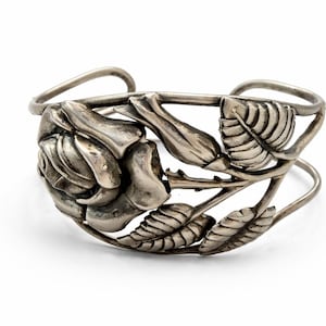 May include: Silver-toned cuff bracelet featuring a detailed rose and leaf design. The floral elements are intricately crafted, with visible petals and veined leaves. The open-ended design allows for easy wear. A decorative piece of jewelry.