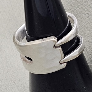 May include: A silver ring featuring a wide, flat, hammered-texture front panel that connects two separate bands. The metal has a polished, reflective finish with visible indentations from the hammering process across the central rectangular section.