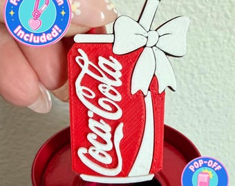 May include: A red Coca-Cola can-shaped charm with white lettering and a white bow. The charm has a straw and is attached to a keychain. The image also includes text that says "Free Keychain Included!" and "POP-OFF pop tabs, not nails."