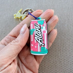 May include: A pink and white striped keychain shaped like a soda can, with the word "Alani" in white script. The keychain has teal accents and a blue loop attached to a silver keyring with a gold glittery charm.