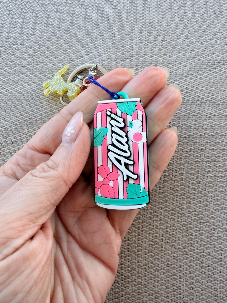 May include: A pink and white striped keychain shaped like a soda can, with the word "Alani" in white script. The keychain has teal accents and a blue loop attached to a silver keyring with a gold glittery charm.