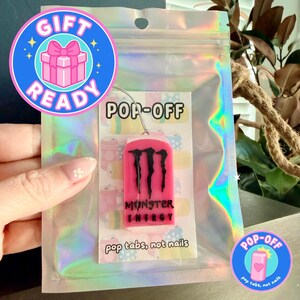 May include: A pink Monster Energy pop-off nail tab with black claw marks and text. The tab is in a clear holographic bag with the text "POP-OFF" and "pop tabs, not nails". A "GIFT READY" sticker is visible.