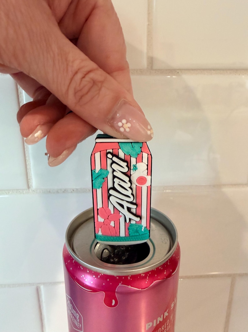 May include: A pink and white striped can-shaped charm with the word "Alani" in white script, adorned with floral and leaf accents. The charm is placed on top of a pink can. The can is open, revealing the interior.