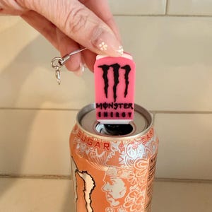 May include: A pink Monster Energy can-shaped charm with the brand's logo and text, attached to a silver chain, sits atop an orange Monster Energy drink can. The can is open, revealing its silver interior.