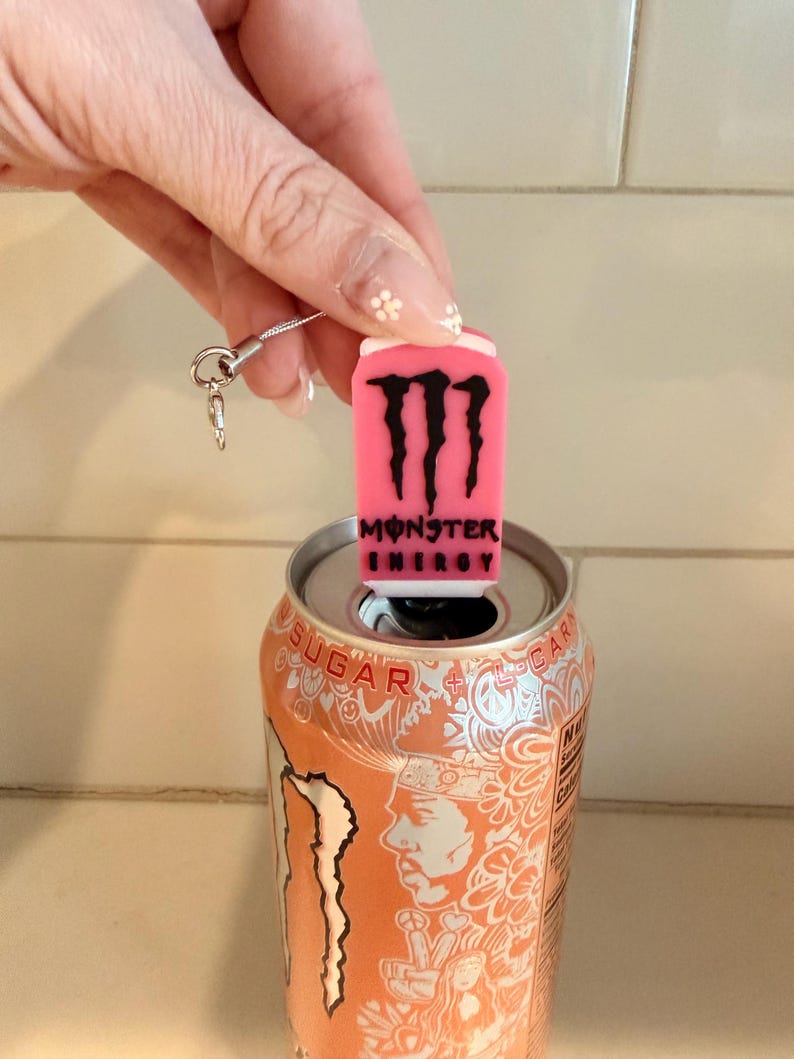 May include: A pink Monster Energy can-shaped charm with the brand's logo and text, attached to a silver chain, sits atop an orange Monster Energy drink can. The can is open, revealing its silver interior.