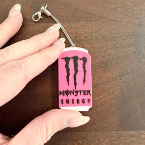 May include: A pink Monster Energy drink can-shaped charm with black lettering and the Monster logo. The charm is attached to a silver-colored lanyard with a lobster clasp.