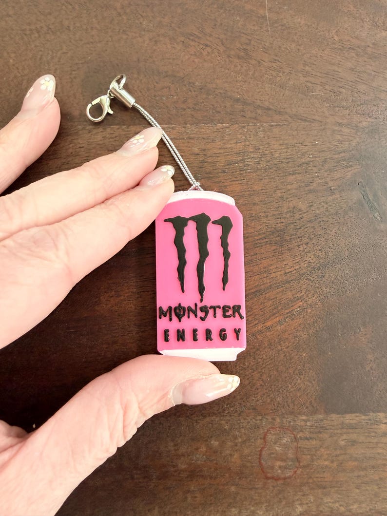 May include: A pink Monster Energy drink can-shaped charm with black lettering and the Monster logo. The charm is attached to a silver-colored lanyard with a lobster clasp.