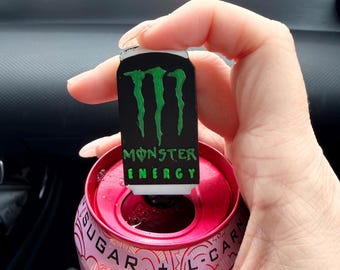 May include: A small rectangular black enamel pin featuring the green Monster Energy logo and text. The pin is positioned over the opening of a pink aluminum beverage can with visible text reading SUGAR + L-CARN on the top rim of the can.