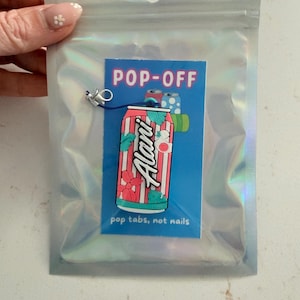 May include: A colorful Alani Nu soda can design on a blue background with the text "POP-OFF" and "pop tabs, not nails." The design is in a holographic bag with a small charm attached.
