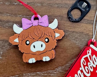 May include: A brown and pink Highland cow charm with a pink bow, white horns, and a red string. The charm is next to a black pull tab and a red Coca-Cola tag.