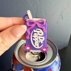 May include: A purple Dr. Pepper can-shaped charm with a white straw and a purple bow. The charm has the Dr. Pepper logo in white and is decorated with small pink flowers. The charm is placed on top of a Dr. Pepper can.